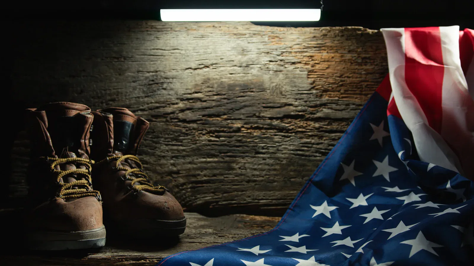 A pair of worn brown combat boots stand on a wooden surface under a bright light. To the right, an folded American flag hangs over the wood, illuminated by the light. The background is a rugged, textured wooden wall, giving the scene a rustic and solemn atmosphere.